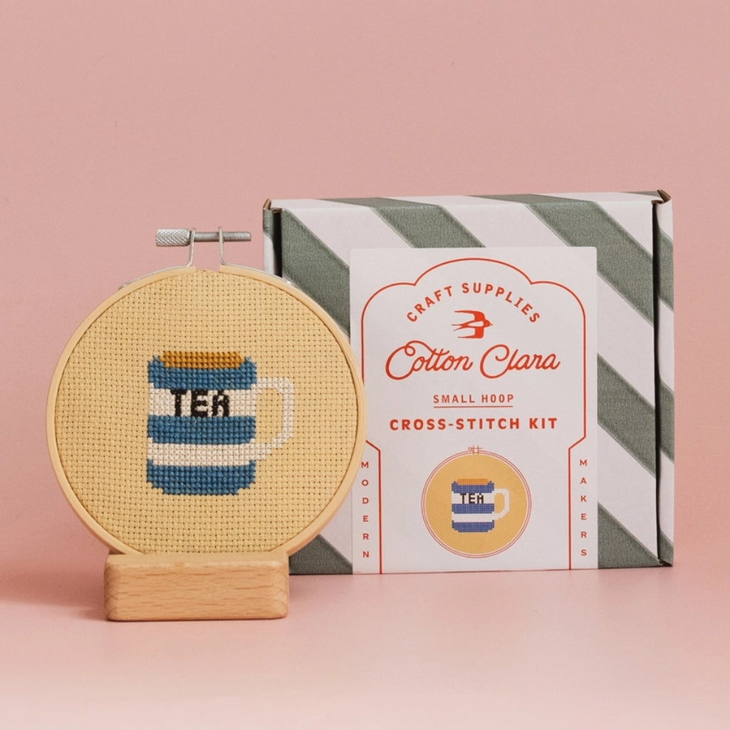 Cross-stitch kit with a completed design of a tea cup and the Cotton Clara packaging on a pink background.