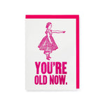 A greetings card featuring a pink illustration of a woman pointing with the words 'You're old now' in bold pink type
