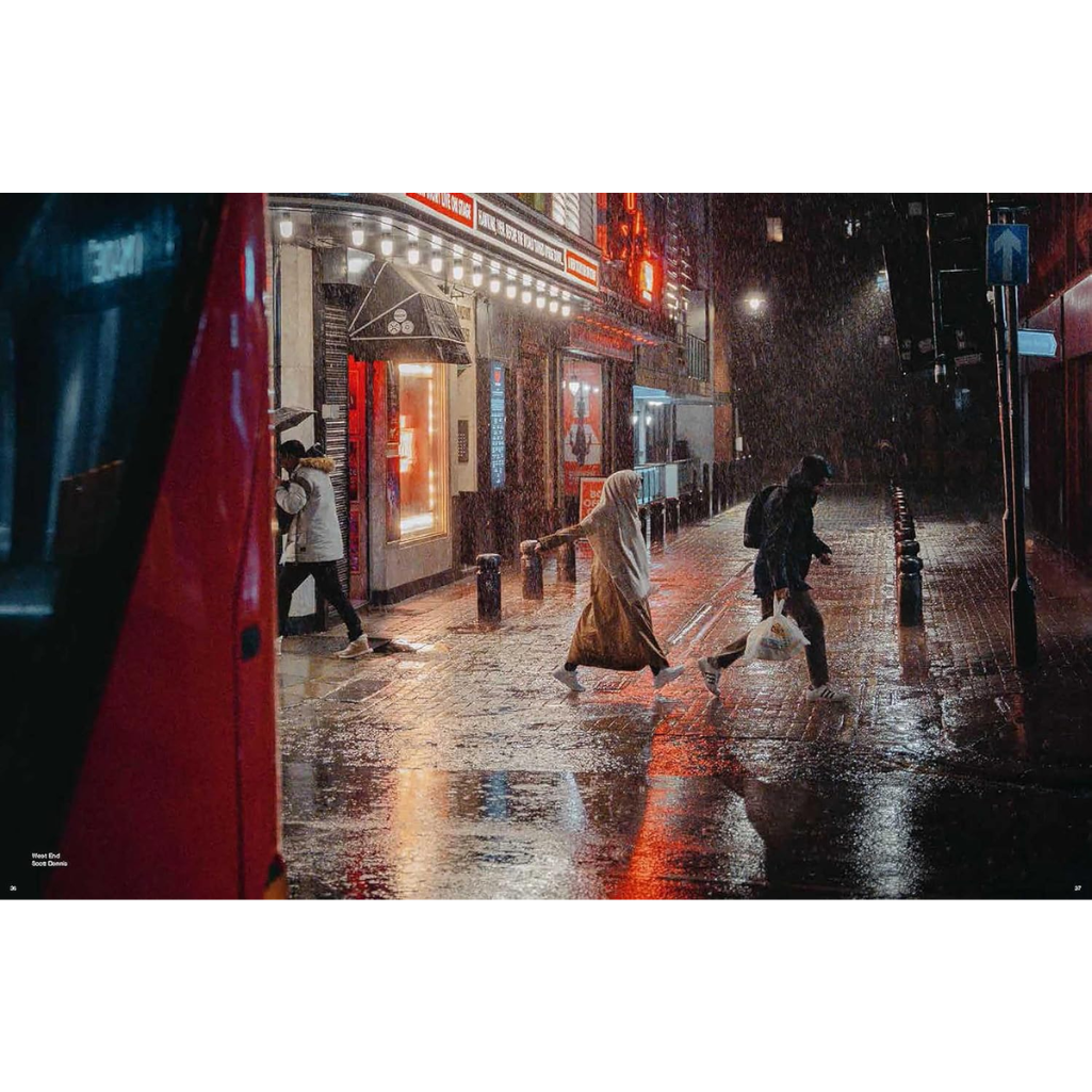 A square art print depicting a rainy city street at night, featuring pedestrians crossing the wet pavement with reflections of storefront lights, framed by a red vehicle on the left.