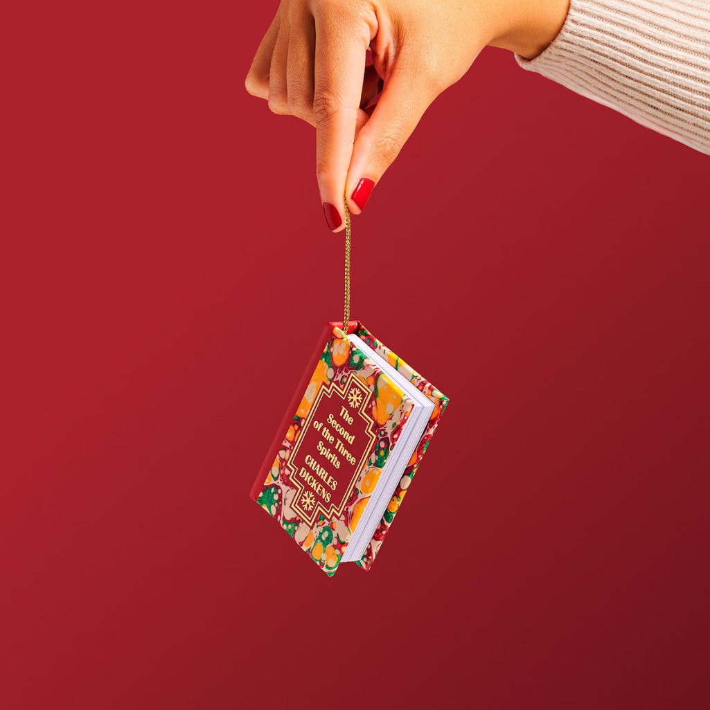 A hand with red nail polish holds a miniature Christmas ornament shaped like a small book with the text 'The Gift of the Christmas Trees' on a solid dark red background.