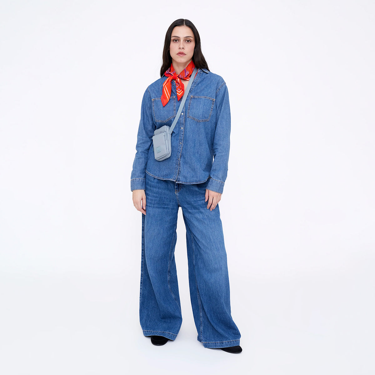 A model stands against a white studio background wearing a blue denim long-sleeve shirt and matching wide-leg jeans, accessorized with a red patterned neck scarf and a small gray crossbody bag.