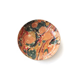 A round ceramic plate with a vibrant marbled paint pattern in orange, red, black, and cream, photographed from directly above on a white background.