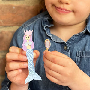 Child holding a paper mermaid figure and a small object with a mirror, against a brick wall background.