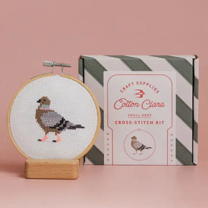 Cross-stitch kit with a completed design of a bird, next to its packaging on a pink background.