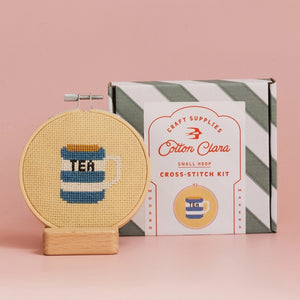 Cross-stitch kit with a completed design of a tea cup and the Cotton Clara packaging on a pink background.
