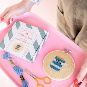 Cross stitch kit with hoop, threads, scissors, and pattern book on a pink tray.